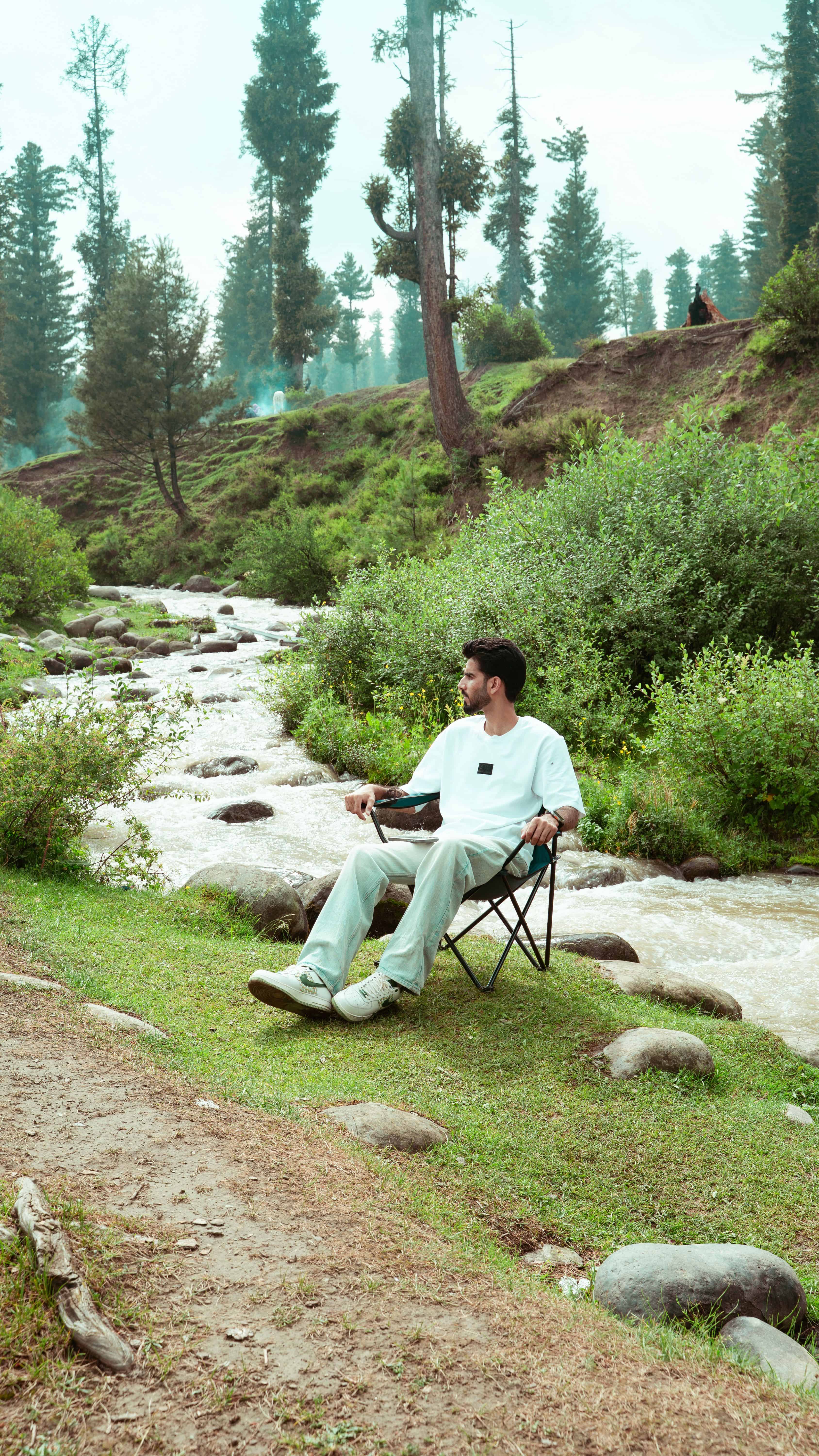 A man sits on a camping chair by a serene mountain stream surrounded by lush greenery in Kashmir. Courtesy: Rubaitul Azad, Pexels